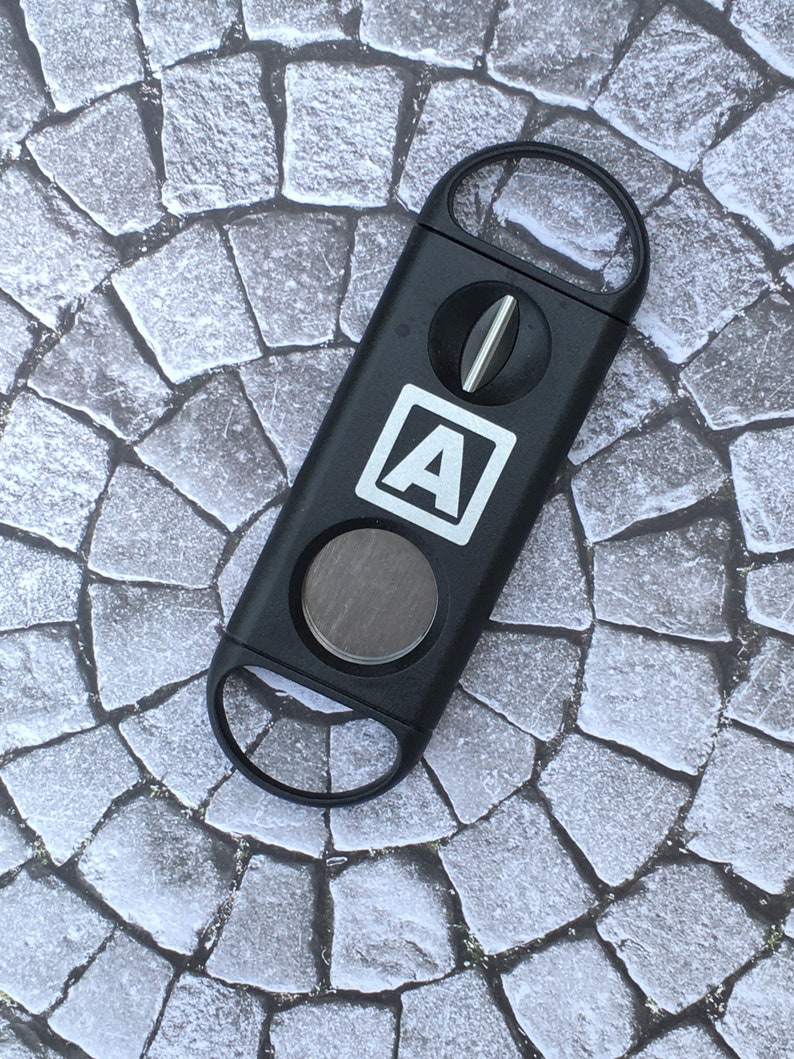 May include: Black cigar cutter with a silver blade and a white "A" logo. The cutter is on a grey stone background.
