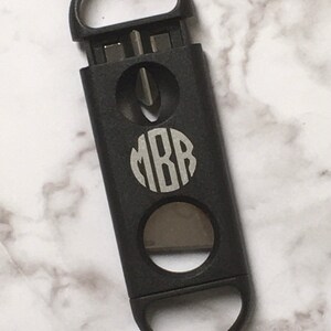 May include: Black cigar cutter with a monogrammed circle that says "MBB" in silver lettering. The cutter has two different sized circular blades.