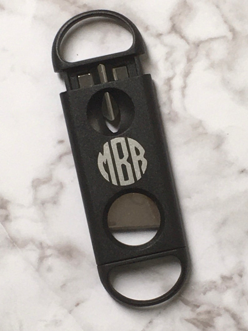May include: Black cigar cutter with a monogrammed circle that says "MBB" in silver lettering. The cutter has two different sized circular blades.