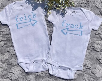 May include: Two white baby onesies with blue text. One onesie says "frick" with an arrow pointing to the right. The other onesie says "frack" with an arrow pointing to the right.