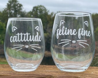 May include: Two clear glass wine glasses with etched cat designs and text. The smaller glass says "cattitude" and the larger glass says "feline fine".