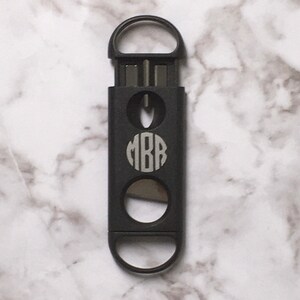 May include: A black cigar cutter with a monogrammed circle that says "MBB" on the front. The cutter has a ring opener on the top and a blade on the bottom.