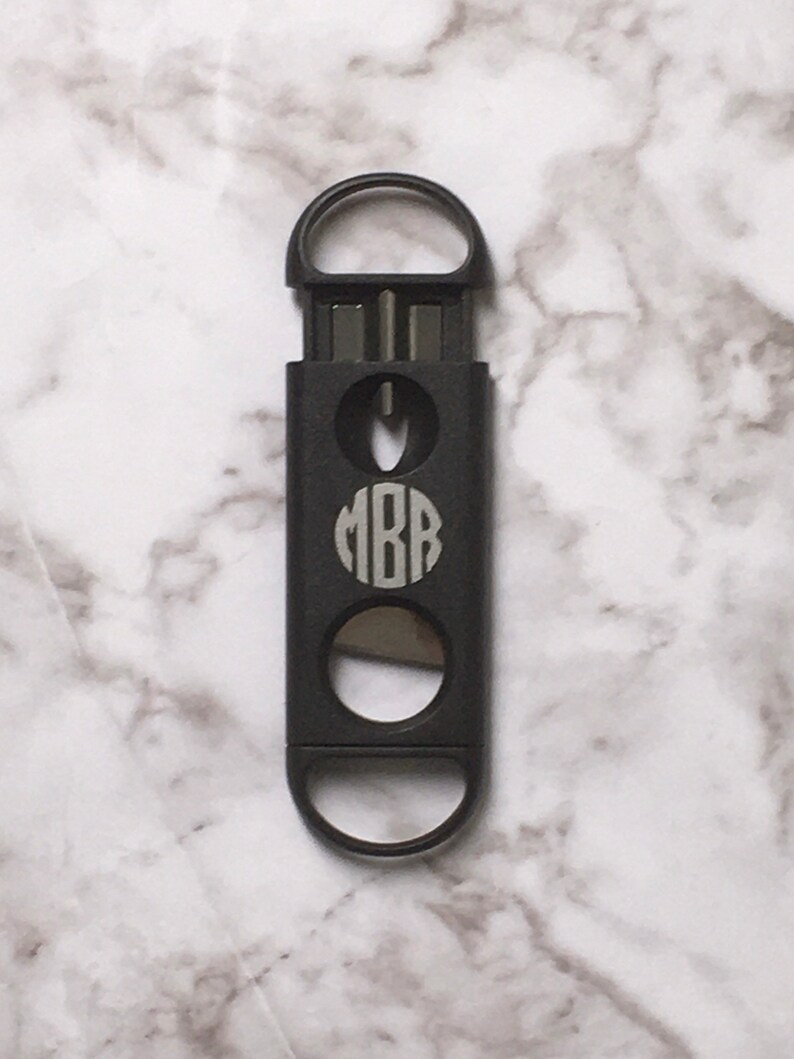 May include: A black cigar cutter with a monogrammed circle that says "MBB" on the front. The cutter has a ring opener on the top and a blade on the bottom.