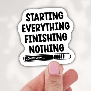 May include: A white sticker with black text that reads "STARTING EVERYTHING FINISHING NOTHING." Below the text is a loading bar with the words "COMING SOON..." The sticker is held by a hand.