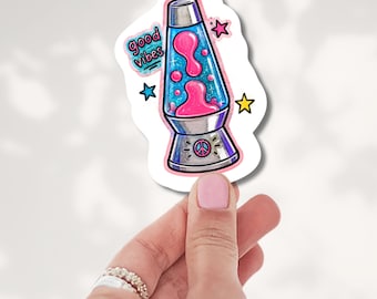 May include: A die-cut sticker featuring a lava lamp with a silver base and cap, containing bright pink liquid blobs inside a blue glittery chamber. The sticker includes the text good vibes, a pink star, a blue star, a yellow star, and a peace sign.