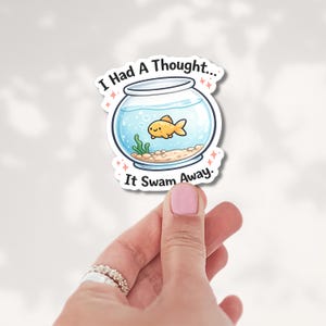 May include: A die-cut sticker featuring a round glass fishbowl with a small orange goldfish, green seaweed, and tan gravel inside. The text "I Had A Thought... It Swam Away." is printed in black above and below the bowl with pink sparkles.
