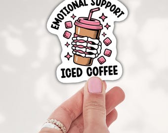 May include: A die-cut sticker featuring a brown iced coffee cup with a pink lid and straw, held by a white skeleton hand. The text reads EMOTIONAL SUPPORT ICED COFFEE in black, surrounded by small pink square ice cubes and star icons.
