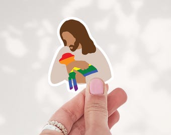 May include: A die-cut sticker featuring a stylized illustration of a figure with long brown hair and a beard, wearing a light beige garment, holding a small lamb colored with horizontal rainbow stripes against a white background.
