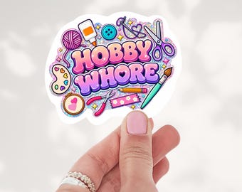 May include: A glittery sticker featuring the text HOBBY WHORE in bold pink and purple gradient letters, surrounded by illustrations of craft supplies including yarn, glue, a button, scissors, a paint palette, a hoop, pliers, a pencil, and tape.