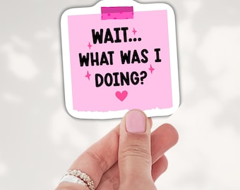 May include: A square pink sticker shaped like a sticky note with a piece of pink tape at the top. The text reads "WAIT... WHAT WAS I DOING?" in black letters, accented by small pink stars and a pink heart at the bottom center.