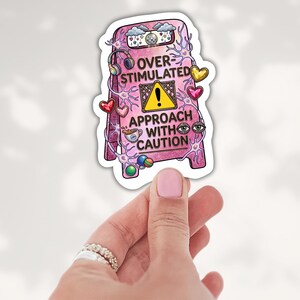 May include: A pink A-frame sign sticker featuring the text OVER-STIMULATED APPROACH WITH CAUTION, a yellow warning triangle, headphones, a disco ball, hearts, neurons, eyes, and a coffee cup, all rendered in a colorful, detailed illustration.