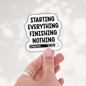May include: A white die-cut sticker with black text that reads STARTING EVERYTHING FINISHING NOTHING. Below the text is a black loading bar graphic with the words COMING SOON... inside, featuring a series of vertical bars indicating progress.