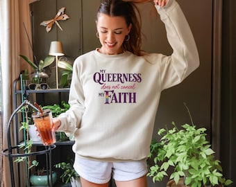 May include: A beige long-sleeve crewneck sweatshirt featuring the purple text "MY QUEERNESS does not cancel MY FAITH," with the word "MY" in the second line stylized with a rainbow-colored gradient pattern across the letters.