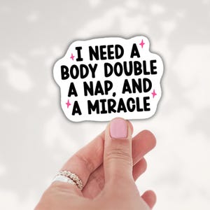 May include: A white die-cut sticker with black text that reads I NEED A BODY DOUBLE A NAP, AND A MIRACLE. The sticker features small pink four-pointed star accents surrounding the text, which is written in a bold, rounded sans-serif font.