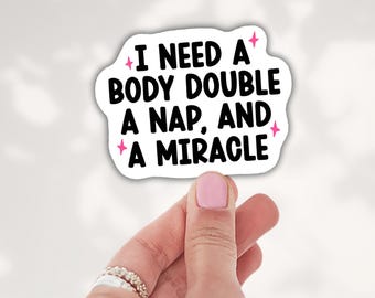 May include: A white die-cut sticker with black text that reads I NEED A BODY DOUBLE A NAP, AND A MIRACLE. The sticker features small pink four-pointed star accents surrounding the text, which is written in a bold, rounded sans-serif font.
