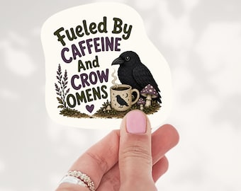 May include: A die-cut sticker featuring a black crow perched next to a steaming mug decorated with a small crow and moon icon. The text reads Fueled By CAFFEINE And CROW OMENS, accompanied by illustrations of purple mushrooms and lavender sprigs.