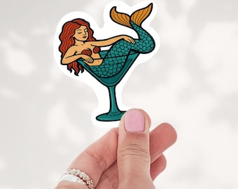 May include: A die-cut sticker featuring a mermaid with long red hair and a teal-scaled tail resting inside a large martini glass. The mermaid has a tan complexion and is positioned with her tail curled upward, rendered in a bold graphic style.