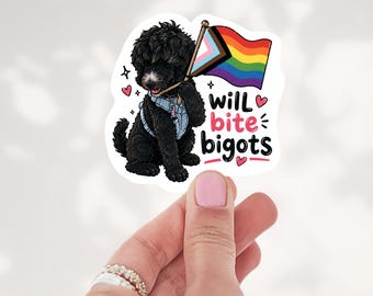 May include: A die-cut sticker featuring a fluffy black dog wearing a light blue harness and holding a flagpole with a rainbow and transgender pride flag. The text reads will bite bigots in black and pink, surrounded by small pink hearts.