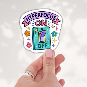 May include: A die-cut sticker featuring a teal light switch with a purple toggle, surrounded by colorful flowers, stars, and hearts. The text reads HYPERFOCUS ON at the top and OFF at the bottom, with a yellow lightning bolt icon.