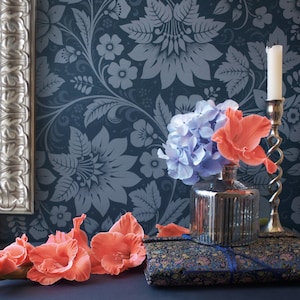 May include: A silver candlestick with a white candle sits on a table with a blue floral patterned wallpaper. A bouquet of blue hydrangeas and pink flowers are in a silver vase on the table.