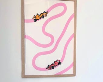 May include: Framed art print featuring two race cars, one orange and one red, racing on a pink track against a white background. The artwork is encased in a light brown wooden frame, suitable for wall decor.