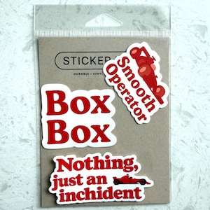 Peut inclure: Un paquet d'autocollants en vinyle avec du texte rouge sur fond blanc. Les autocollants incluent les phrases "Box Box", "Smooth Operator" et "Nothing, just an inchident". Un graphique de voiture de course rouge est également inclus.