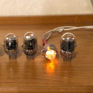 May include: Four clear glass vacuum tubes with silver bases and internal components. One tube glows with a bright orange light. The tubes are connected by wires and sit on a wooden surface.