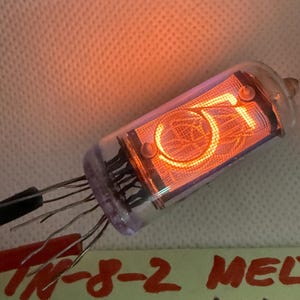 May include: A vintage TN-8-2 vacuum tube with a glowing orange internal display. The glass tube has multiple wire leads extending from the base. The text "TN-8-2 MEL" is visible on a yellow sticky note.