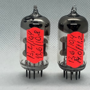 May include: Two vintage vacuum tubes with black metal tops and clear glass bodies. The tubes have a red sticker with handwritten numbers on them. The numbers on the left tube are 131/104 and 136/108. The numbers on the right tube are 136/104 and 140/112.