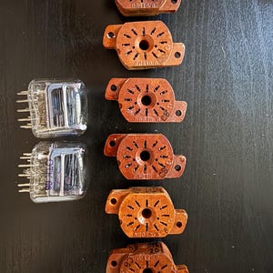 Pode incluir: Dois tubos de vácuo de vidro transparente com pinos metálicos e estruturas internas de arame, rotulados OTK 66, ao lado de seis soquetes eletrônicos circulares de baquelite marrom. Cada soquete possui um orifício central e doze ranhuras.