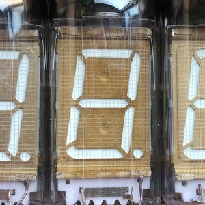 Pode incluir: Três tubos Nixie vintage com uma caixa de vidro transparente, cada um exibindo o número "8" em um formato digital segmentado branco. Os tubos estão colocados sobre um fundo marrom.