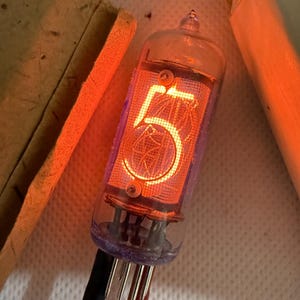 May include: A vintage Nixie tube displaying the number 5 in a warm orange glow. The glass tube has a purple base and multiple pins. Black and red wires are attached. The background includes a cardboard box and a textured surface.