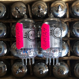 May include: A close-up of a cardboard box filled with vintage vacuum tubes. Two clear glass tubes with pink labels are in focus, displaying handwritten numbers. The tubes have silver bases and internal components, with the text "6Ж32П" and "EX77" visible.
