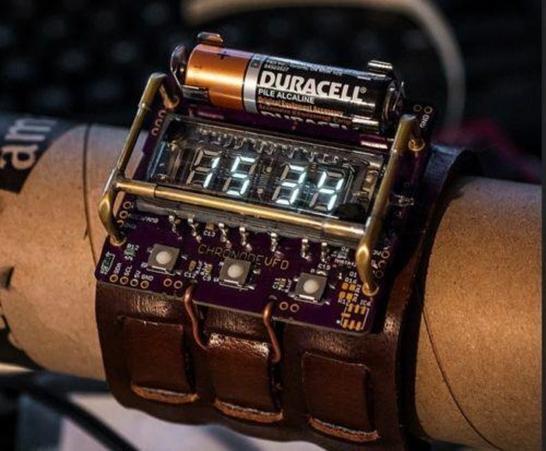 K&ouml;nnte beinhalten: Eine Steampunk-Armbanduhr mit digitalem Display, einem Lederarmband und einer Duracell-Batterie, die an der Seite befestigt ist. Das Zifferblatt der Uhr zeigt "088:88" an und die Platine ist mit "CHRONODE UFD" beschriftet.