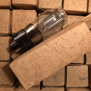 May include: A glass vacuum tube with a black base and internal elements, partially out of its cardboard box. The box rests on a stack of similar light brown boxes, each with handwritten markings. The tube is a vintage electronic component.