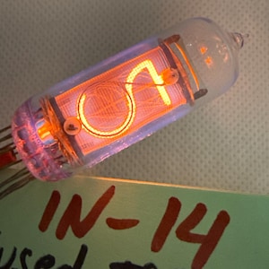 May include: A clear glass IN-14 vacuum tube with an orange glowing display. The display shows a stylized number "A" with a circular element. The tube is labeled "IN-14 used, tested".