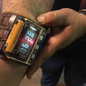 May include: A brown leather wristband with a silver buckle and a digital watch face with a red and green display. The watch face reads "12:21".
