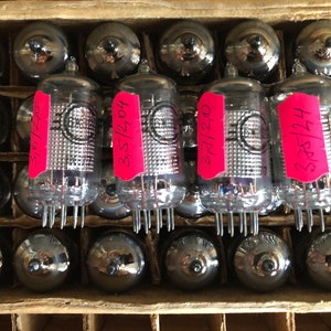 May include: A close-up of four vintage vacuum tubes in a cardboard box. The tubes are labeled with numbers and have a silver finish. The box is brown and has a grid pattern.