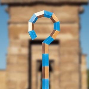 May include: A blue and gold papercraft staff with a curved top, resembling an ancient Egyptian ankh symbol. The staff is in focus, while the background is blurred, showing a stone structure.