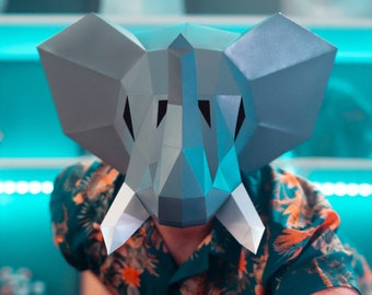 May include: A person wearing a gray paper mache elephant mask with white tusks. The mask is geometric in shape and has a blue and gray color scheme. The person is wearing a green and orange patterned shirt.