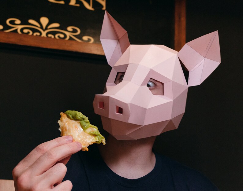 K&ouml;nnte beinhalten: Eine Person mit einer rosafarbenen Papier-Schweinemaske h&auml;lt ein dreieckiges St&uuml;ck Essen, das mit gr&uuml;nem Guacamole belegt ist.