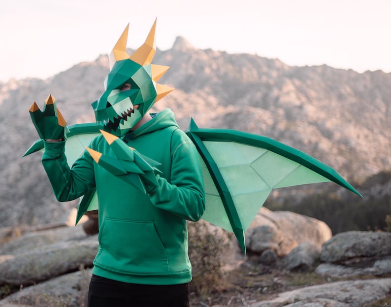 May include: A person wearing a green dragon costume with a paper mache mask, wings, and claws. The costume is detailed and realistic, with sharp teeth and claws. The person is standing in a mountainous landscape.