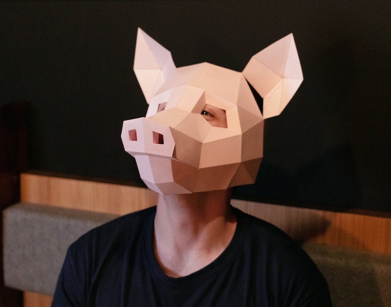 K&ouml;nnte beinhalten: Eine Person tr&auml;gt eine rosafarbene Papier-Schweinemaske. Die Maske besteht aus gefaltetem Papier und hat einen 3D-Effekt. Die Person tr&auml;gt ein schwarzes Hemd.
