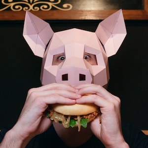 K&ouml;nnte beinhalten: Eine Person mit einer rosafarbenen Schweinemaske isst einen Hamburger. Der Hamburger hat Salat, Zwiebeln und einen Patty.