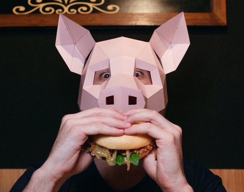 K&ouml;nnte beinhalten: Eine Person mit einer rosafarbenen Schweinemaske isst einen Hamburger. Der Hamburger hat Salat, Zwiebeln und einen Patty.
