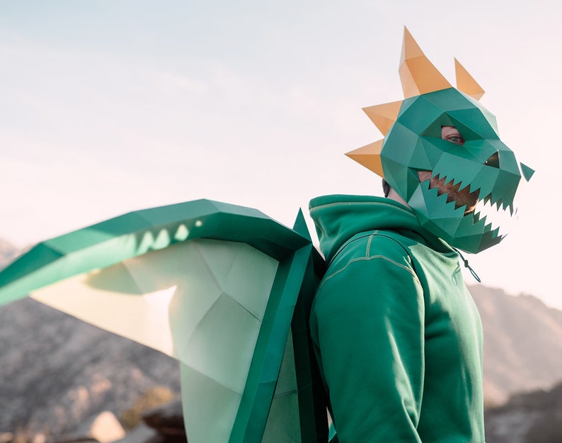 May include: A person wearing a green hoodie and a green and gold paper dragon mask with wings. The mask has a sharp, toothy grin.