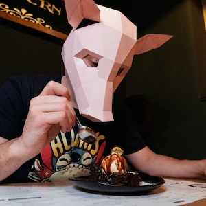 K&ouml;nnte beinhalten: Eine Person mit einer rosa Schweinemaske isst ein Schokoladen-Dessert mit Eis und Schokoladensauce. Das Dessert befindet sich auf einem schwarzen Teller und die Person h&auml;lt einen L&ouml;ffel.