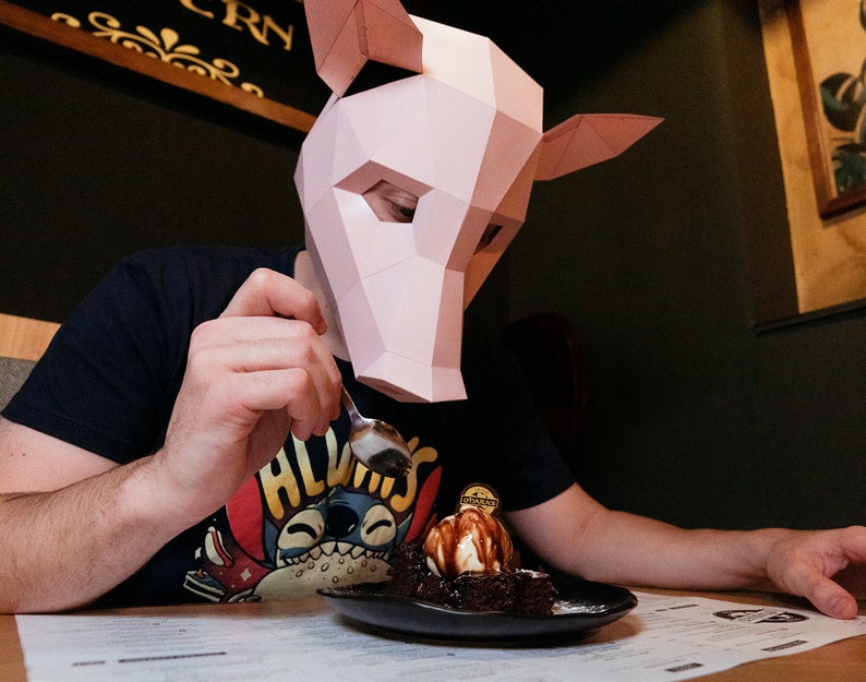 K&ouml;nnte beinhalten: Eine Person mit einer rosa Schweinemaske isst ein Schokoladen-Dessert mit Eis und Schokoladensauce. Das Dessert befindet sich auf einem schwarzen Teller und die Person h&auml;lt einen L&ouml;ffel.