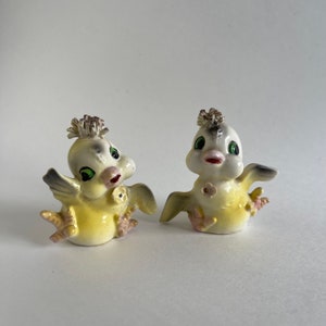 May include: Two ceramic yellow chicks with green eyes and pink beaks. They are wearing crowns and have a flower on their chests. They are both facing forward and have their wings out.