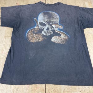 Può includere: Una t-shirt nera a maniche corte con una stampa grafica di un teschio umano con un serpente a sonagli marrone avvolto attorno. Il testo "Austin 3:16" è stampato in un carattere scuro sulla fronte del teschio, circondato da un bagliore blu.